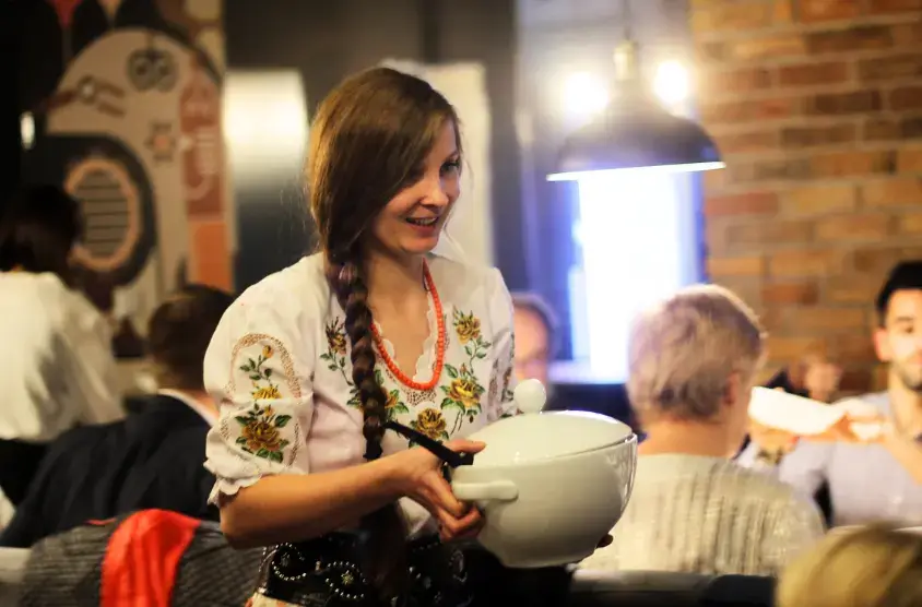 Uśmiechnięta kelnerka w stroju góralskim podaje danie gościom podczas wigilii w górach.