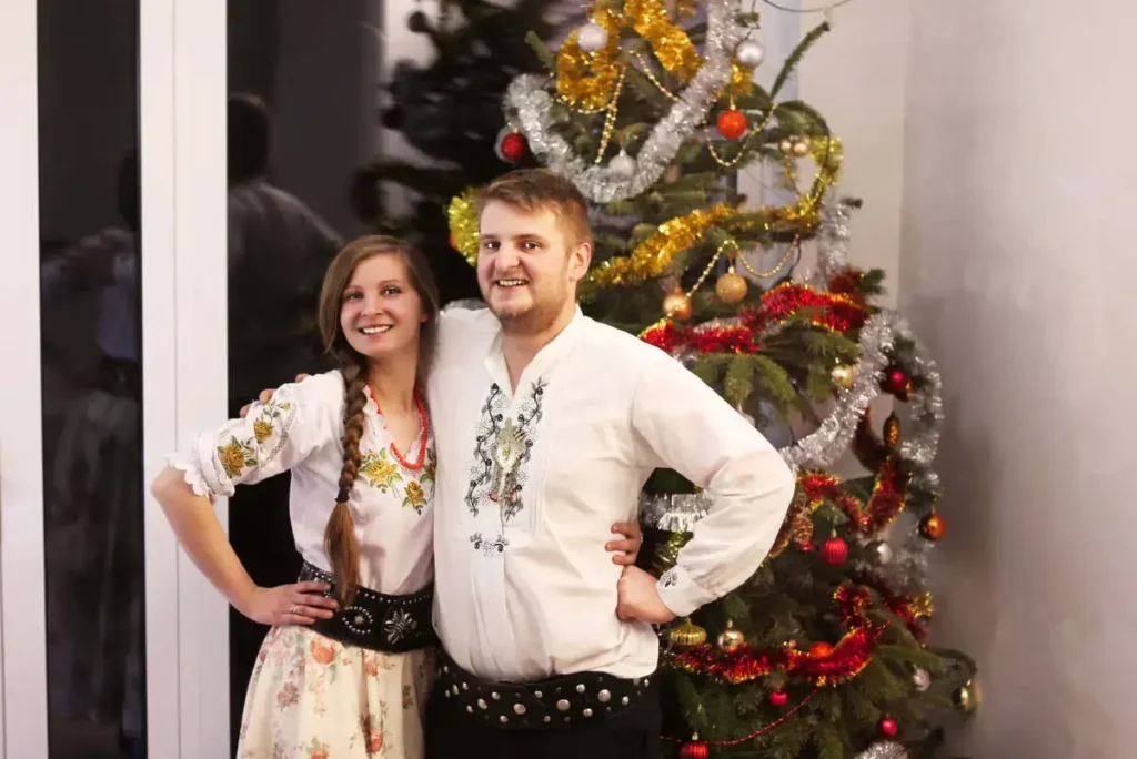 Uśmiechnięta para w strojach regionalnych na tle choinki, oferująca pakiety bożonarodzeniowe w górach.